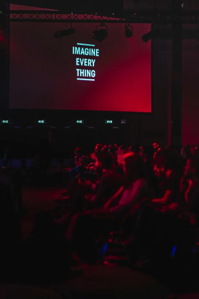 Corporate conference audience watching a keynote on a large screen in a red-lit venue—professional event photography.